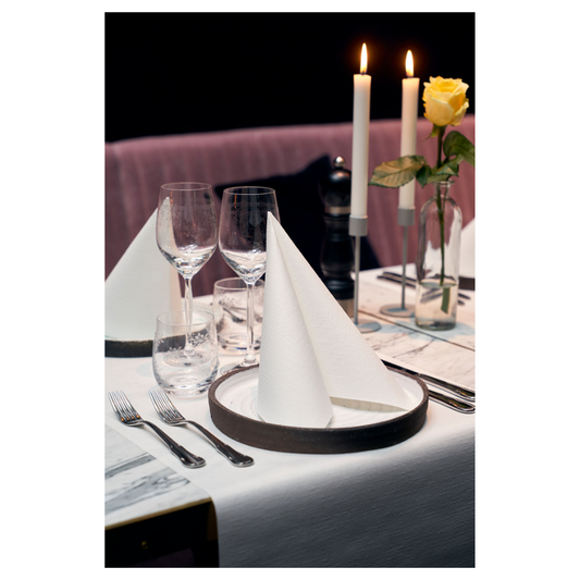 Ein Esstisch ist elegant gedeckt mit zwei Duni Dunilin®-Servietten 48 x 48 cm Weiß, die zu Dreiecken gefaltet auf Tellern liegen. Zwei große brennende Kerzen und eine gelbe Rose in einer Glasvase zieren den Hintergrund. Weingläser, ein Wasserglas und Besteck vervollständigen dieses anspruchsvolle Ambiente mit stoffähnlichem Charme.