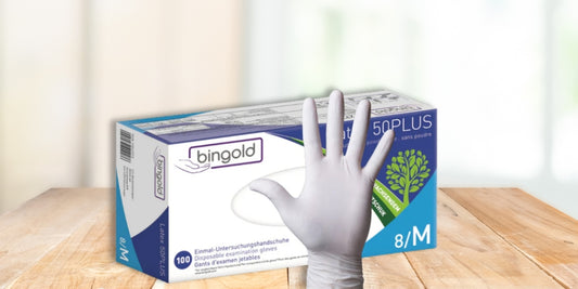 Eine Schachtel mit Bingold 50 Plus Einweg-Untersuchungshandschuhen, Größe Medium, mit einer Hand, die einen weißen Handschuh trägt, vor der Schachtel, auf einer Holzfläche.