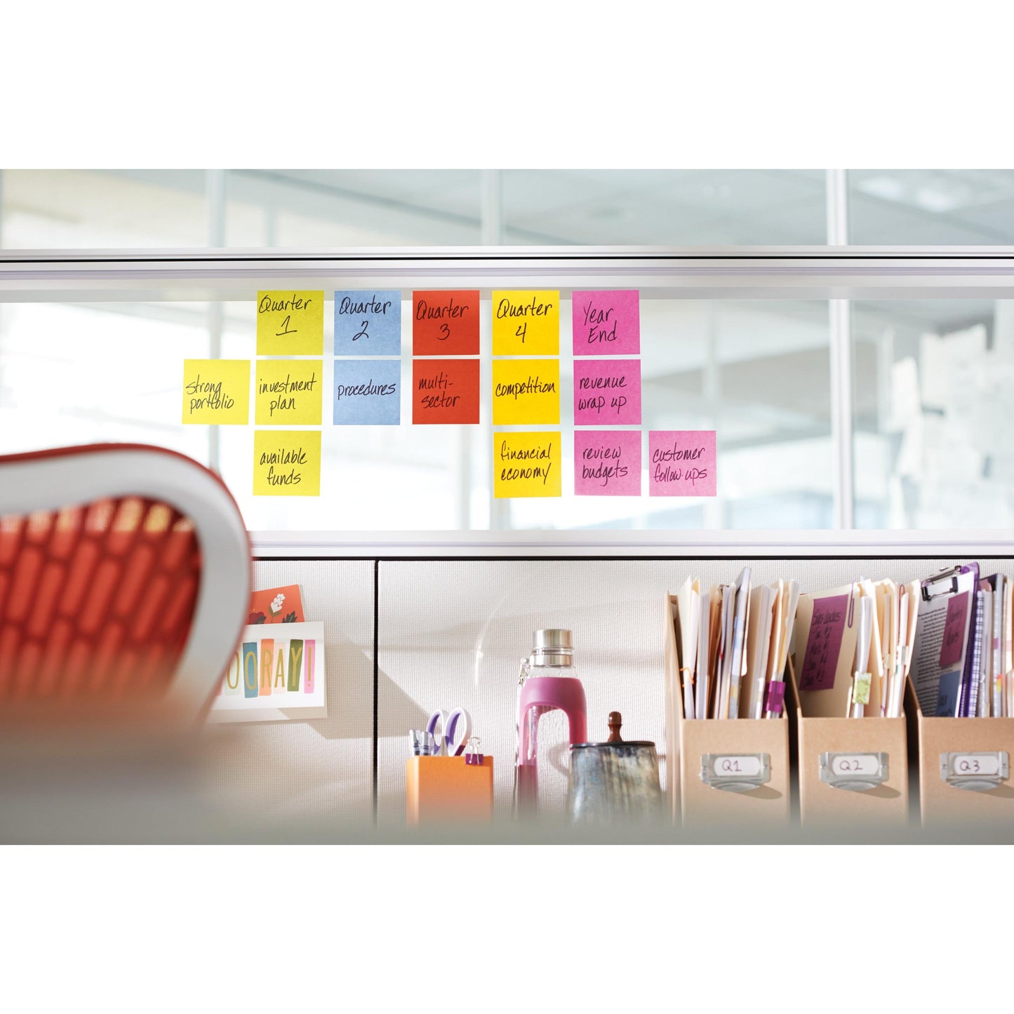 Im Büro zeigt eine Trennwand die vierteljährlichen Aufgaben auf bunten 3M Post-it® Super Sticky Z-Notes (76 x 76 mm, 20+4 Blöcke) an, davor stehen ein oranger Stuhl und Regale mit Flaschen, Ordnern und Mappen.