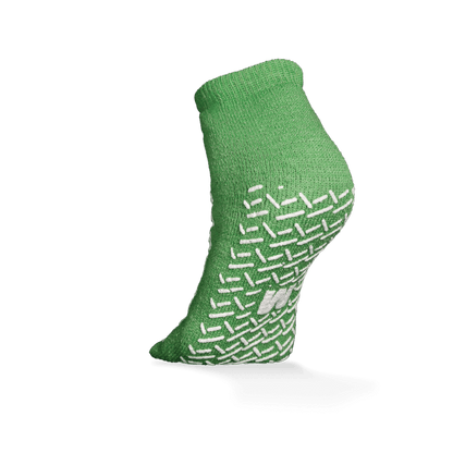 The ECO-PLUS anti-slip disposable socks, double-sided coated by AMPri Handelsgesellschaft mbH, is a green ankle sock with white non-slip grips, depicted on an elevated foot against a white background - ideal for medical areas.