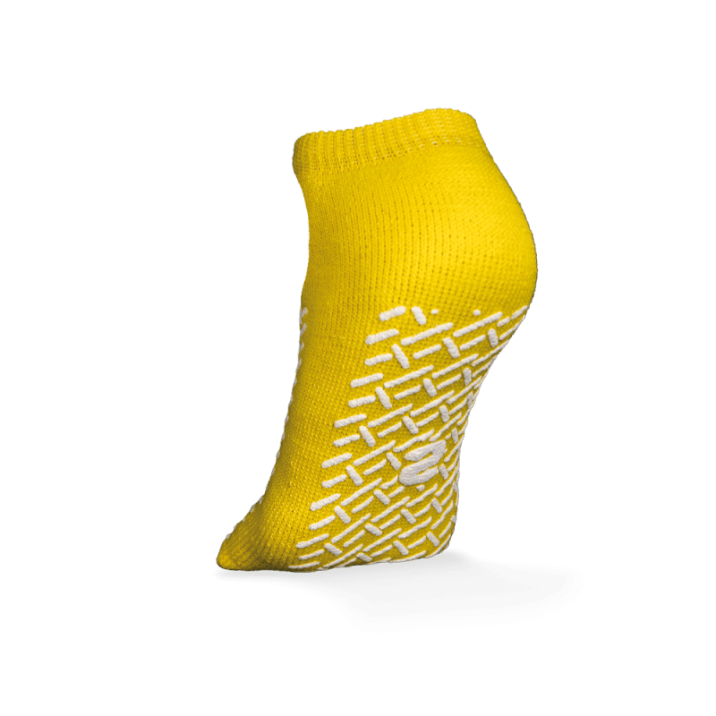 A single yellow ECO-PLUS anti-slip disposable sock, double-sided coated by AMPri Handelsgesellschaft mbH, stands upright on a white background, with white geometric anti-slip grips and a central logo, ideal for medical areas.