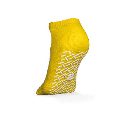 A single yellow ECO-PLUS anti-slip disposable sock, double-sided coated by AMPri Handelsgesellschaft mbH, stands upright on a white background, with white geometric anti-slip grips and a central logo, ideal for medical areas.