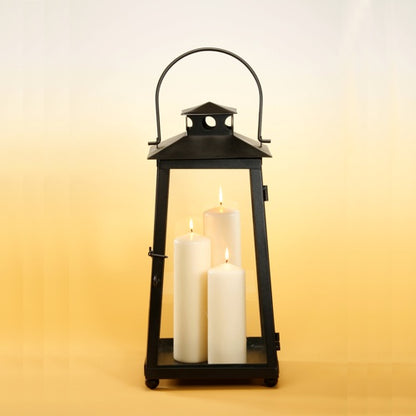 A black metal lantern with a handle contains three burning Starpak 3 fireplace and lantern candles Ø 70 mm ivory (pack, 3 pieces), against a gradient background from light yellow at the bottom to pale yellow at the top.