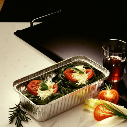 A Starpak 25 tray aluminum dish (PP-coated with insert lid) is filled with uncooked green pasta nests, tomato slices, basil, and grated cheese on a white surface. Next to it are a glass with a red drink, whole tomatoes, and leafy greens.
