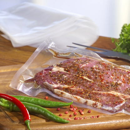 A Starpak 100 vacuum bag, PA/PE transparent, filled with marinated meat lies on a wooden cutting board amidst chili peppers and pink peppercorns. Leafy greens and utensils complete the scene - ideal for use with a chamber vacuum sealer.