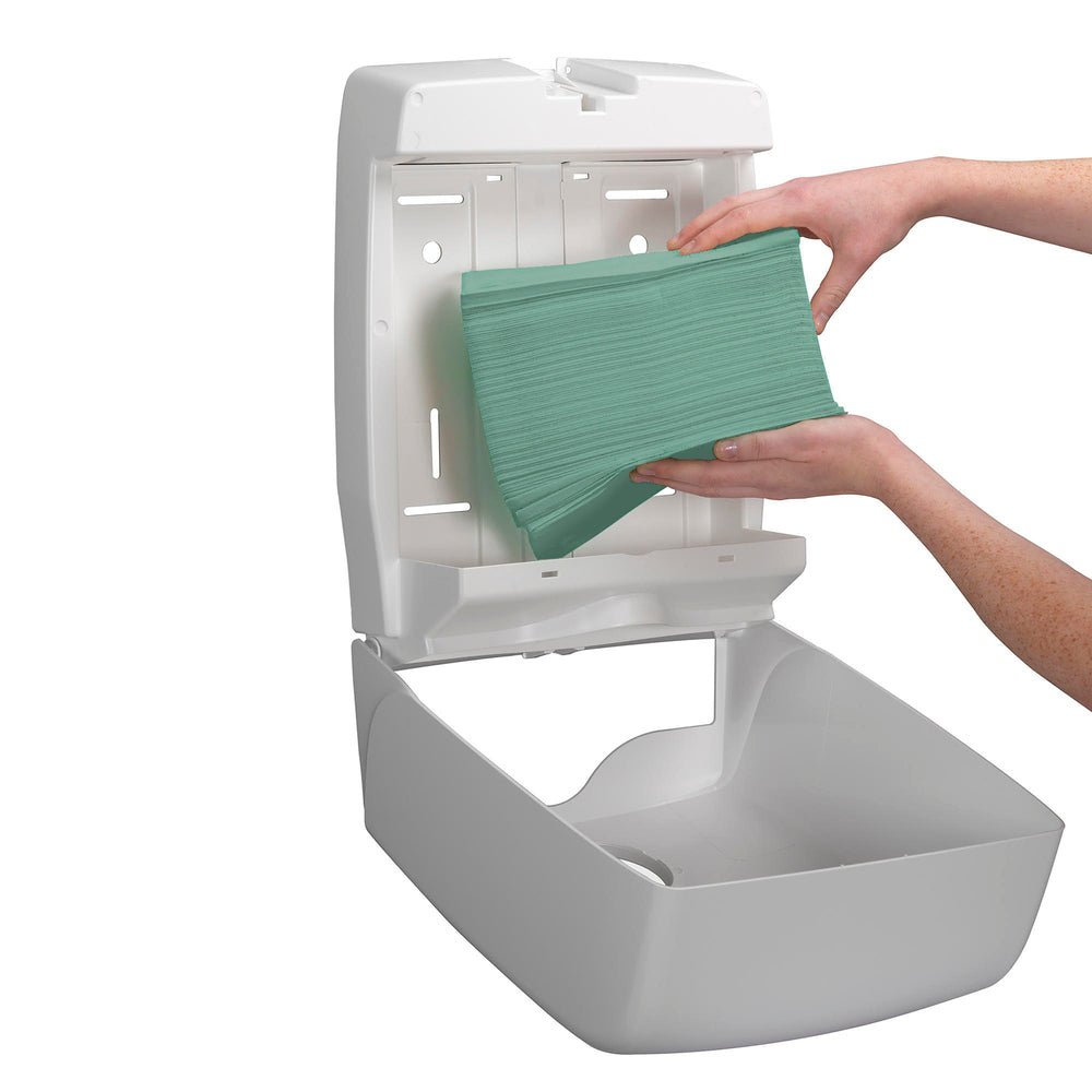 Eine Person legt einen Stapel Kimberly-Clark GmbH Hostess™ NATURA™ Handtücher - Zick-Zack-Faltung, Klein (23,00 cm x 25,00 cm) in einen offenen weißen, an der Wand befestigten Papierhandtuchspender.