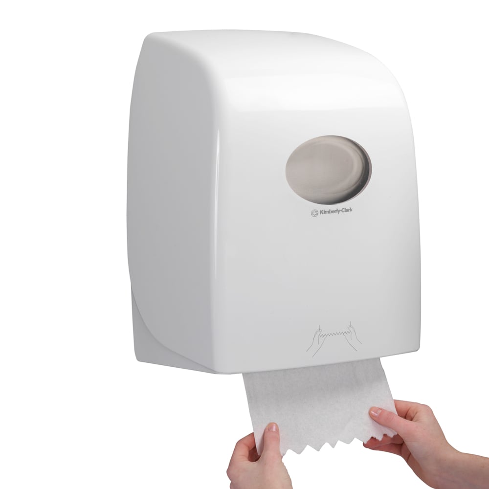 Someone pulls a sheet of Scott® ESSENTIAL™ towel roll from a white, wall-mounted dispenser by Kimberly-Clark GmbH, whose transparent window reveals the towel rolls inside.
