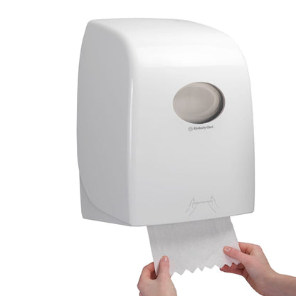 Someone pulls a sheet of Scott® ESSENTIAL™ towel roll from a white, wall-mounted dispenser by Kimberly-Clark GmbH, whose transparent window reveals the towel rolls inside.