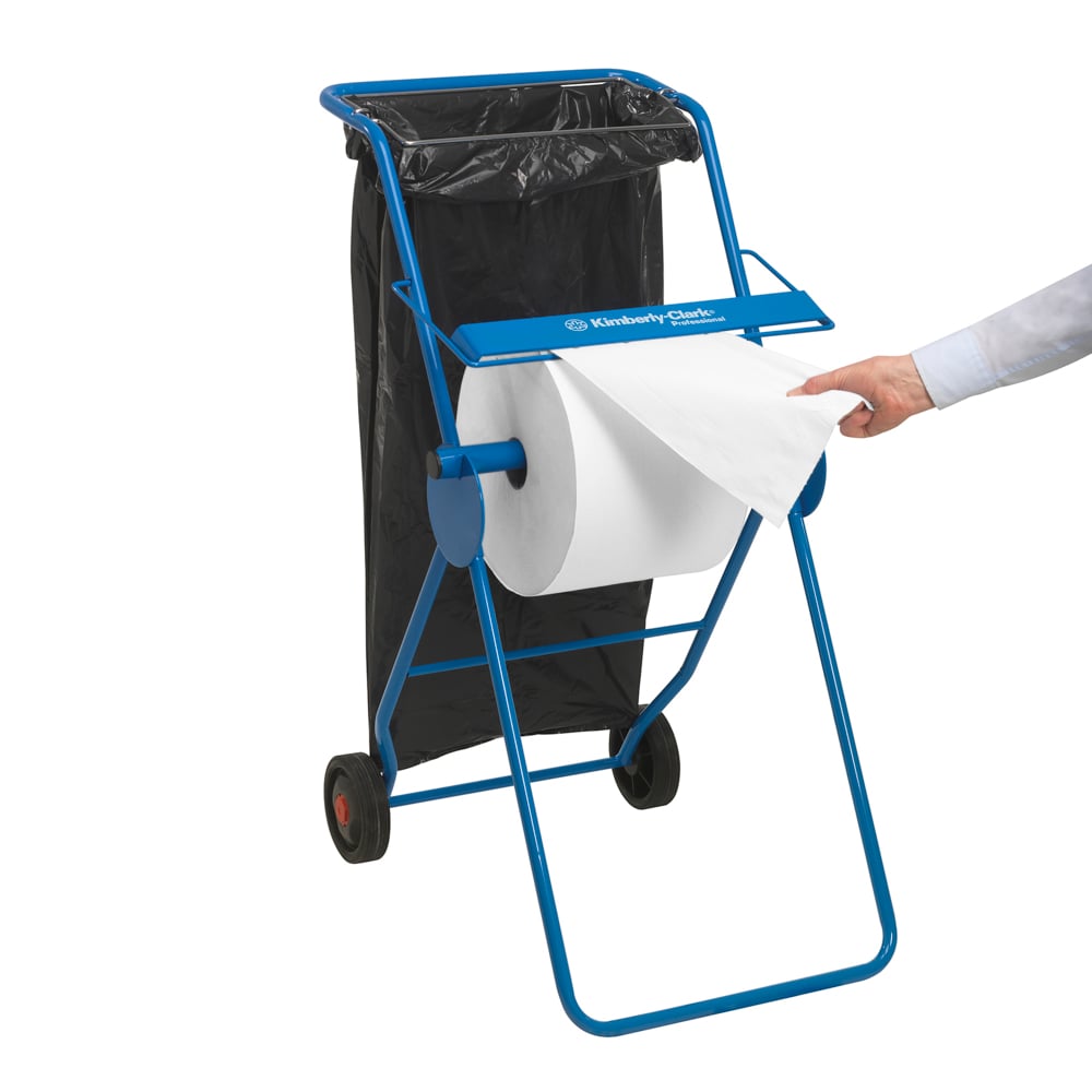 Ein blauer Papierhandtuchspender mit einer großen weißen Scott® ESSENTIAL™ Ärzterolle (6 x 200 Blatt) der Kimberly-Clark GmbH unterstreicht die Hygiene; ein schwarzer Müllsack ist in der Nähe, während die Hand einer Person ein Blatt von der Rolle zieht.