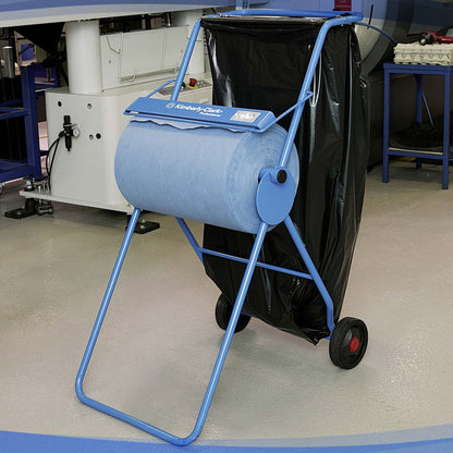 A Kimberly-Clark Professional™ Mobile Stand for Wiper Rolls (Large Roll/Blue) with a roll of blue industrial paper towels and a large black garbage bag stands on wheels on a gray hall floor at a workplace.