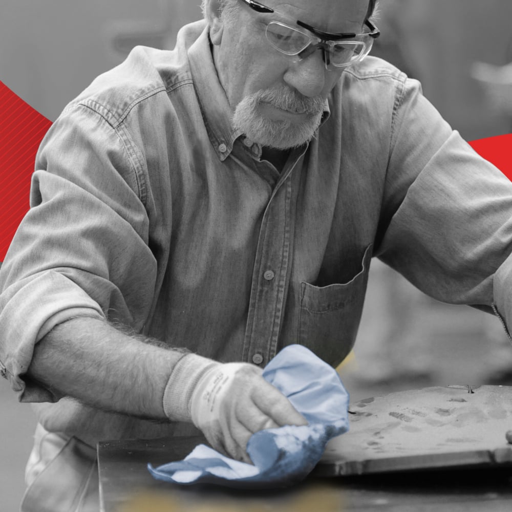 Ein Mann mit Schutzbrille und Handschuhen benutzt ein WypAll® L20 Papierwischtuch für Instandhaltungsarbeiten - Großrolle / Blau von Kimberly-Clark GmbH, um eine Metalloberfläche zu reinigen. Er trägt ein zugeknöpftes Hemd, mit verschwommenen roten und gelben Akzenten im Hintergrund.