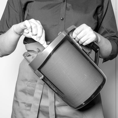 A person in an apron and buttoned shirt stirs a large jug while a WypAll® Reach™ Centrefeed Dispenser (Black) from Kimberly-Clark GmbH stands nearby, adding a professional touch. The black-and-white image highlights the scene.