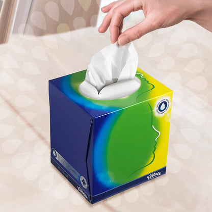 A hand pulls a tissue from the Kleenex® facial tissues - cube box / white from Kimberly-Clark GmbH with blue, green, and yellow design and a white oval opening. A stylized profile suggests facial care in front of a gently blurred background.