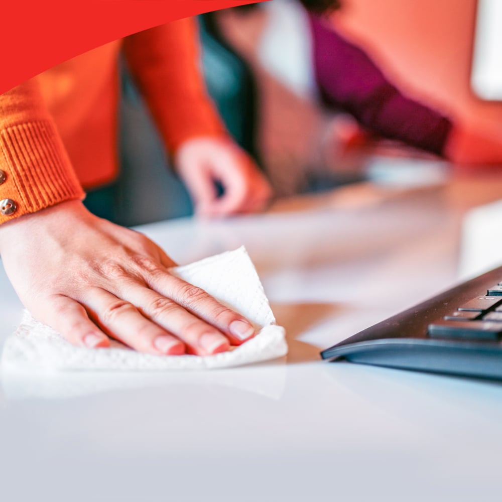 Eine Person in einem orangefarbenen Pullover reinigt einen weißen Schreibtisch in der Nähe einer schwarzen Tastatur mit WypAll® X60 General Clean weißen viertelgefalteten Tüchern (12 Beutel x 76 Blatt) der Kimberly-Clark GmbH, wobei eine andere Person im Hintergrund unscharf zu sehen ist.