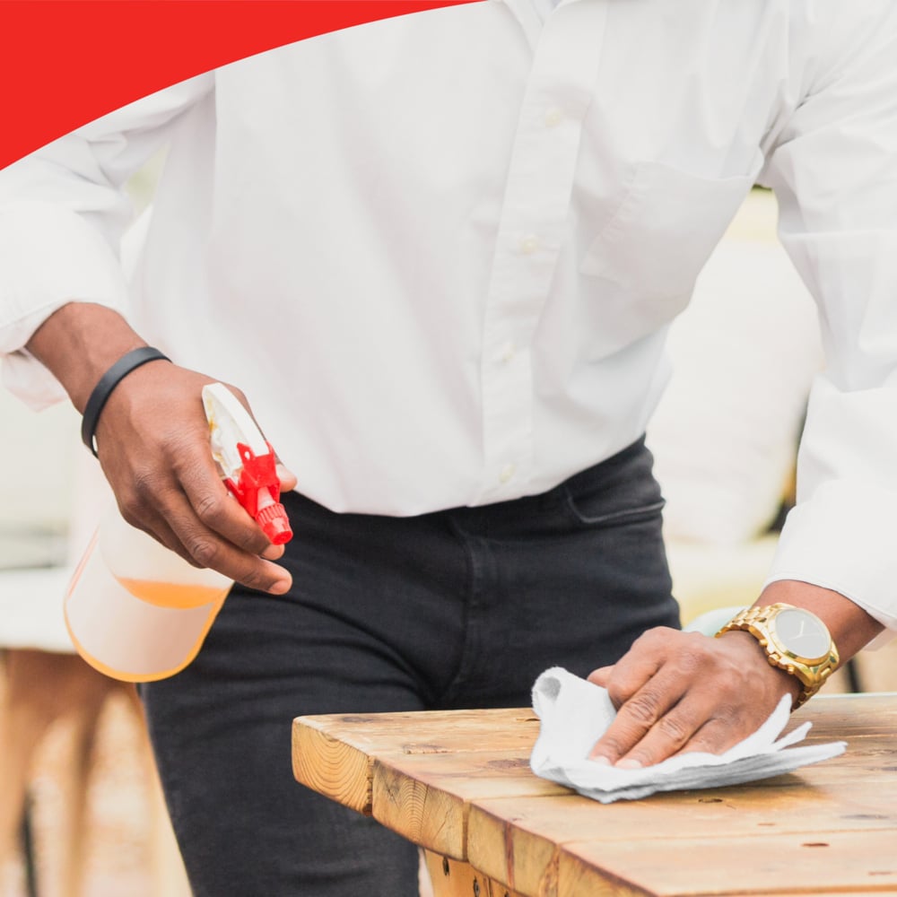 Eine Person in weißem Hemd und schwarzer Hose reinigt einen Holztisch mit WypAll® X60 General Clean Tüchern von Kimberly-Clark GmbH und hält eine Sprühflasche. Die Szene zeigt effiziente Reinigung und einen roten geschwungenen Rand am oberen Rand.