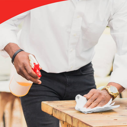 Eine Person in weißem Hemd und schwarzer Hose reinigt einen Holztisch mit WypAll® X60 General Clean Tüchern von Kimberly-Clark GmbH und hält eine Sprühflasche. Die Szene zeigt effiziente Reinigung und einen roten geschwungenen Rand am oberen Rand.