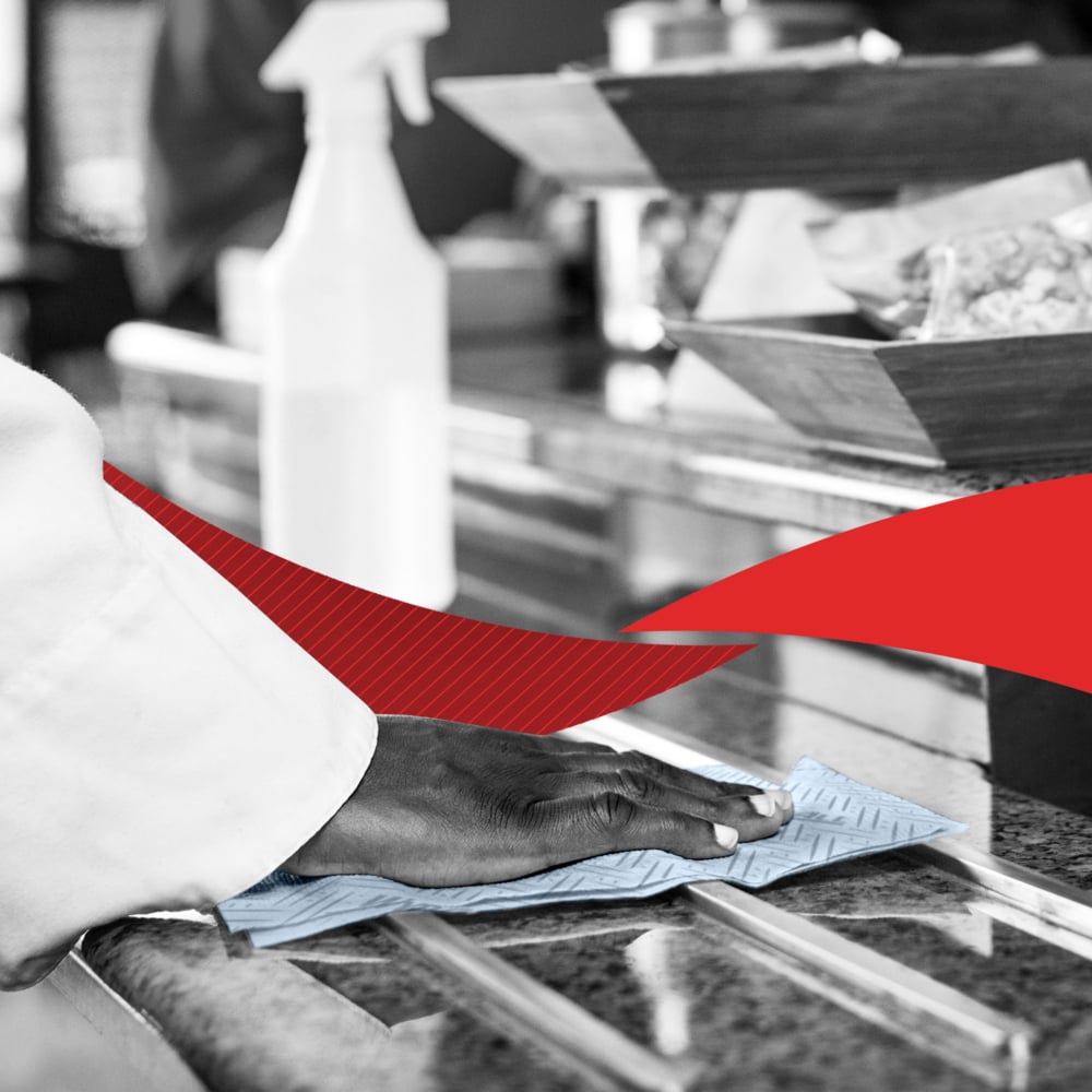 A person wipes a counter with a blue cloth showing cleaning. On the counter are a WypAll® L20 General Clean - Centrefeed for Reach+™ roll and trays from Kimberly-Clark GmbH with a red graphic element in the middle of the image.