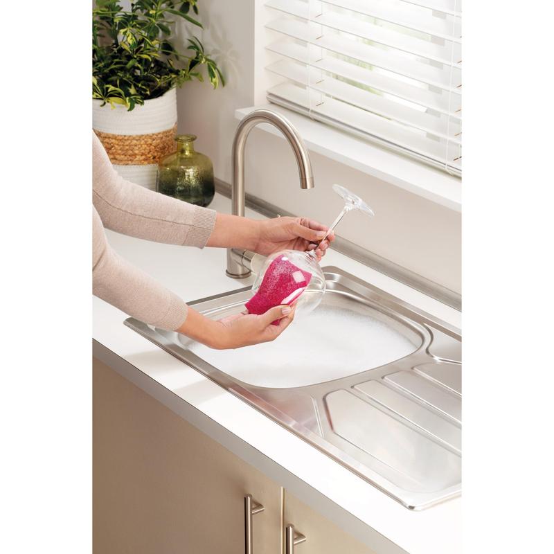 A person cleans a stemmed wine glass with a Scotch-Brite® Delicate Color non-scratch cleaning sponge (2 pieces per pack) from 3M Deutschland GmbH over a soapy sink. Nearby are a potted plant and a green jug.