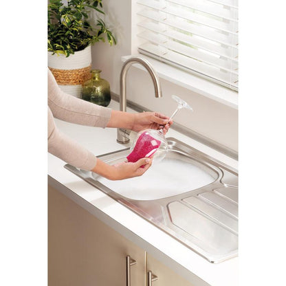 A person cleans a stemmed wine glass with a Scotch-Brite® Delicate Color non-scratch cleaning sponge (2 pieces per pack) from 3M Deutschland GmbH over a soapy sink. Nearby are a potted plant and a green jug.