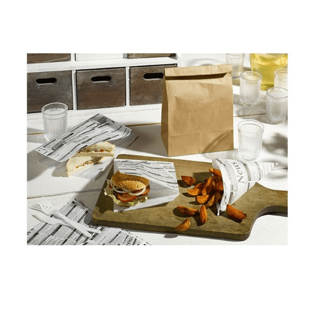 A sandwich and potato wedges are served on a wooden board, and PAPSTAR GmbH "Newsprint" hamburger bags, grease-proof, 16x18 cm (pack of 300), used as a substitute for parchment paper, along with disposable cutlery, napkins, water glasses, and a brown paper bag.
