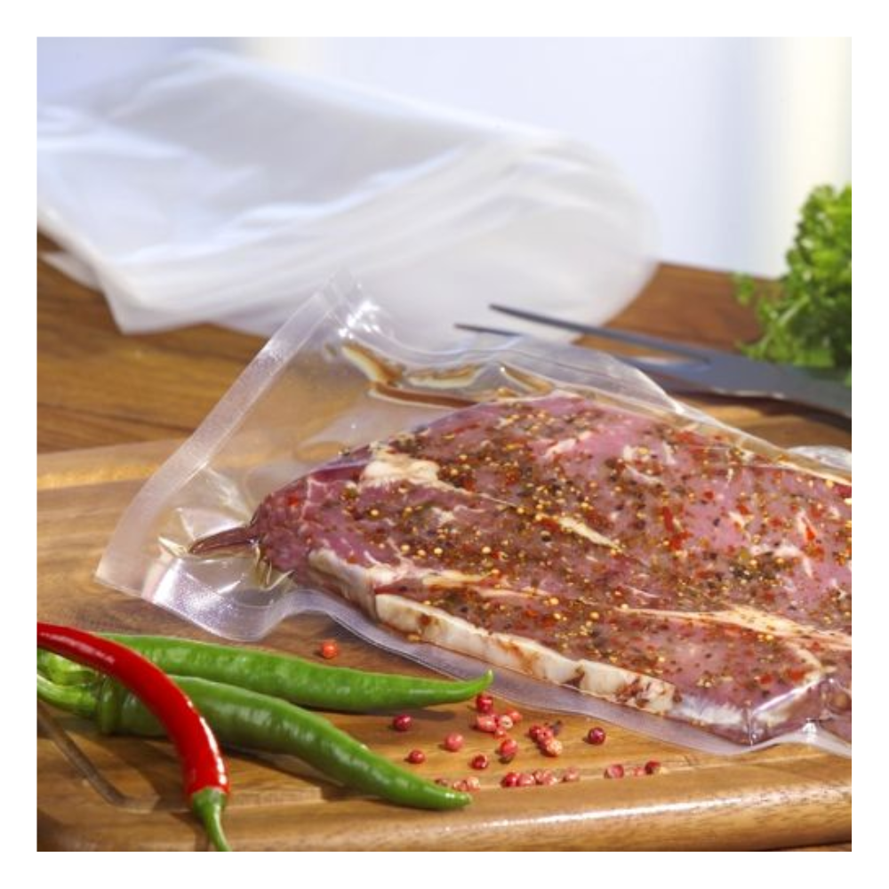 A raw, marinated steak is vacuumed in a transparent Starpak 100 vacuum bag (PA/PE) with a chamber vacuum sealer on a wooden board, with chili peppers, pink peppercorns, leafy greens, and a fork in the background.