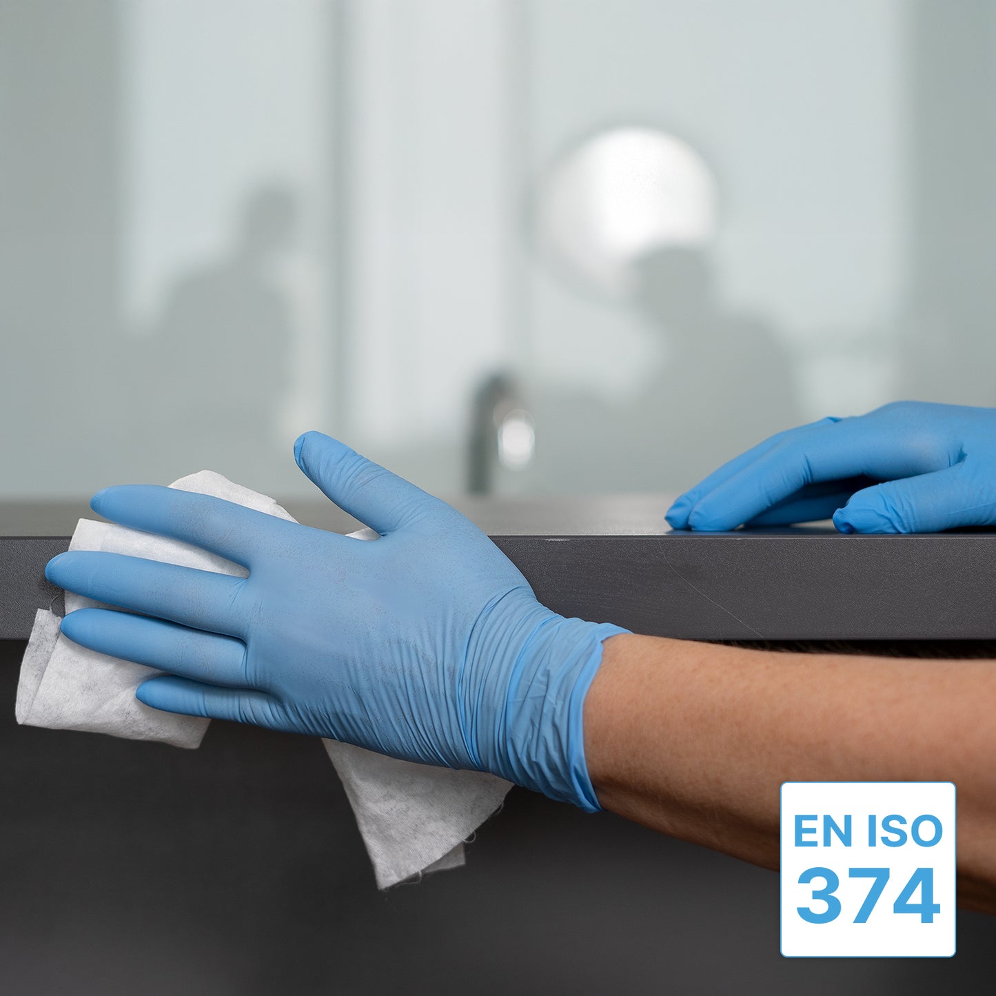 A person wearing ARNOMED NITRIL BLUE XTRA STRONG disposable gloves from ARNOWA GmbH is wiping down a dark countertop with a white cloth. The EN ISO 374 symbol is visible, and an office setting appears blurred in the background.