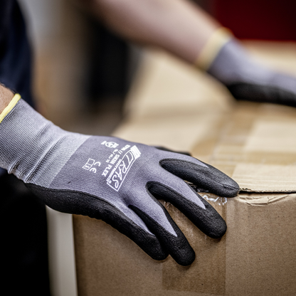 A person wearing NITRAS SKIN FLEX knit gloves from AS Arbeitsschutz GmbH made of special stretch knit, gray, partially coated, lifts a sealed box. Only the gloved hands and part of the box are visible.