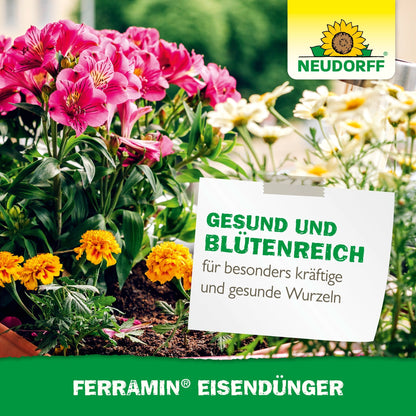 A garden bed with pink and yellow blooms bears a German sign promoting healthy growth and strong roots, with the Ferramin iron fertilizer brand mark from Neudorff Shop, known for combating nitrogen deficiency.