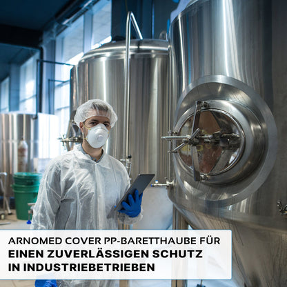 Eine Person in Schutzkleidung, darunter die ARNOMED COVER PP-BARETTHAUBE der ARNOWA GmbH und eine Maske, hält eine Tablette neben Edelstahltanks in einem hygienischen Industriebereich. Der deutsche Text über zuverlässigen Schutz ist unten eingeblendet.