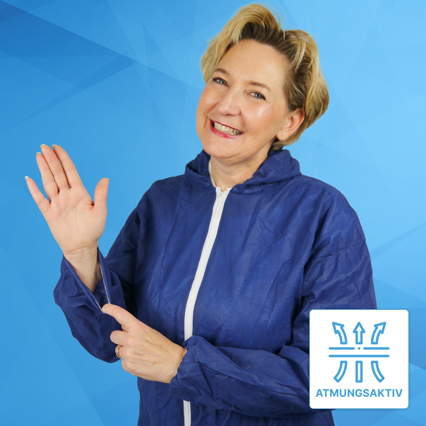Eine Frau im ARNOMED COVER PP-OVERALL BLUE Einmaloverall mit Kapuze aus PP-Vlies von ARNOWA GmbH lächelt und zeigt auf den Ärmel, auf dem ein "ATMUNGSAKTIV"-Symbol das atmungsaktive Gewebe vor einem blauen geometrischen Hintergrund hervorhebt.