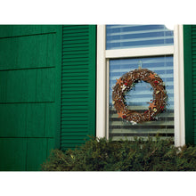 Ein Zweigkranz mit Blättern und kleinen Ornamenten hängt in einem Fenster mit weißen Jalousien, gesichert durch einen transparenten, mittelgroßen Command™ Haken M für den Außenbereich (oval, 17091AW) von 3M Deutschland GmbH. Grüne Fensterläden rahmen das Fenster ein, davor stehen Büsche.