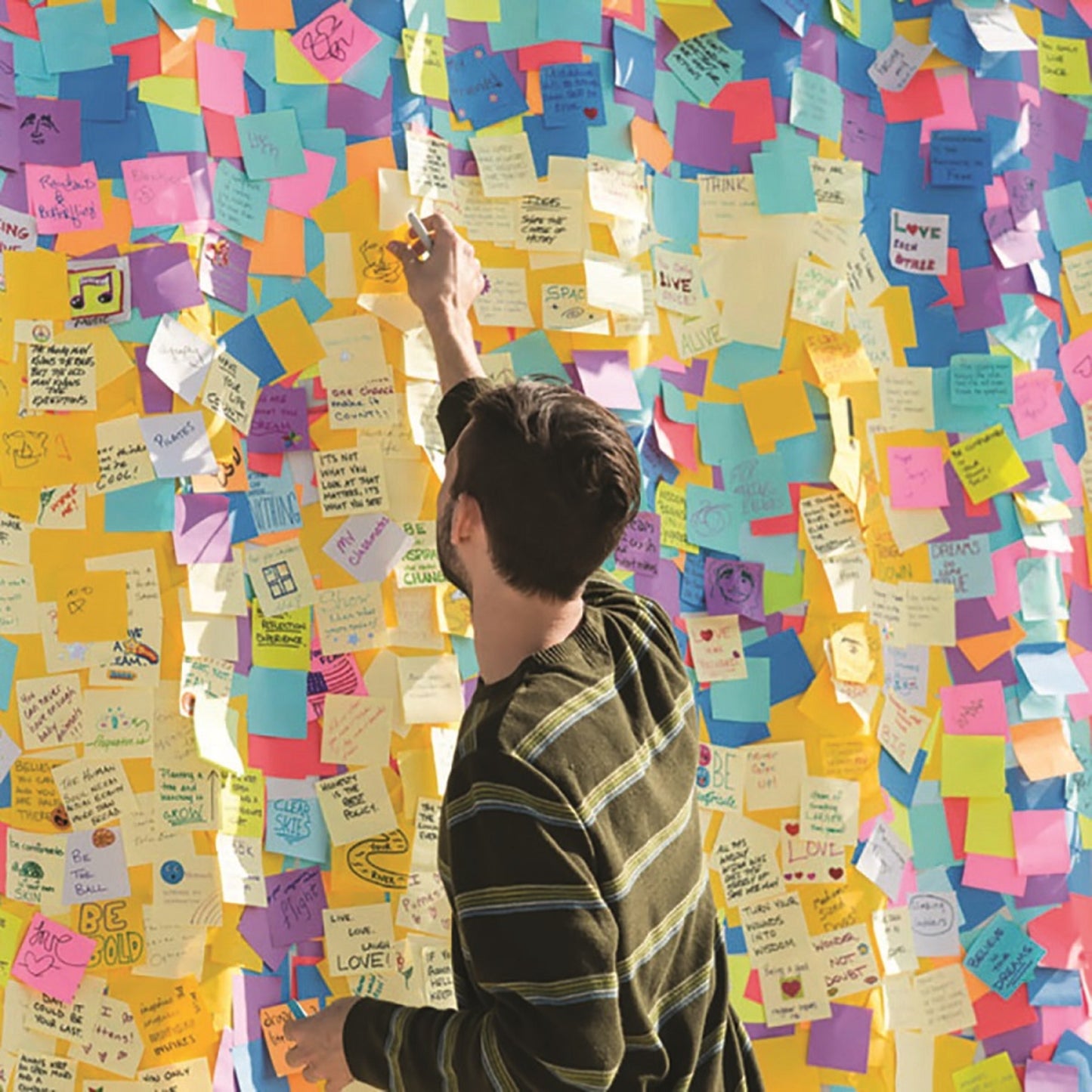 Eine Person im gestreiften Hemd steht an einer Wand mit handgeschriebenen Nachrichten und Zeichnungen und greift nach einem 3M Deutschland GmbH Post-it® Notes Würfel in Ultra-Farben (76x76 mm, 450 Blatt, 100% PEFC), um ihn zwischen die bunten Zettel zu legen.