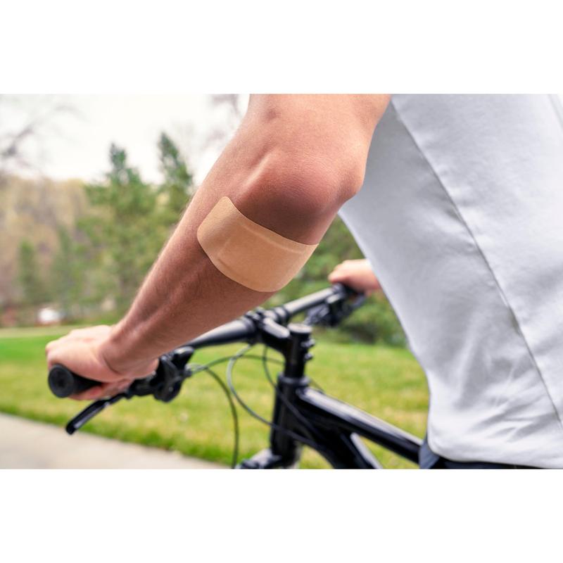 Eine Person, die ein weißes Hemd trägt und im Freien Fahrrad fährt, hat ein Nexcare™ Ultra Stretch MAXI Comfort Flexible Pflaster (50 mm x 101 mm) von 3M Deutschland GmbH auf dem nackten Oberarm; im Hintergrund sind grünes Gras und Bäume zu sehen.
