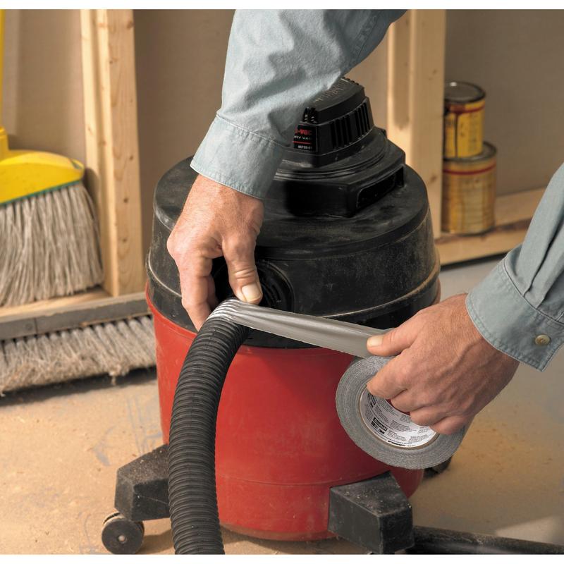 Eine Person, die ein langärmeliges Hemd trägt, verwendet 3M Deutschland GmbH Scotch® Universal Klebeband 2904 (25 m x 48 mm), um den Schlauch eines rot-schwarzen Werkstattsaugers zu befestigen. Im Hintergrund sind ein Besen und Farbdosen zu sehen.