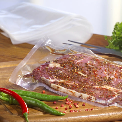 A Starpak 100 vacuum seal bag, PA/PE transparent from Starpak with marinated raw meat lies on a wooden cutting board, surrounded by red and green chili peppers and leafy greens in the background.