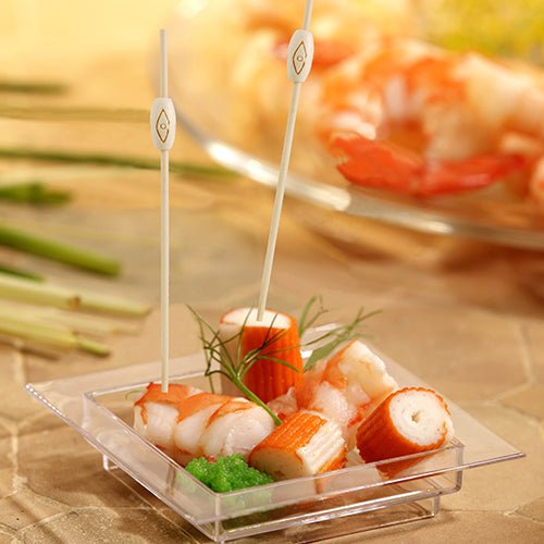 Auf einer quadratischen Kunststoffschale mit Krabbenimitat, Garnelen, grünem Rogen und Garnitur sind zwei PAPSTAR 40 Fingerfood - Picker „pure“ der PAPSTAR GmbH abgebildet, im Hintergrund ist unscharf ein ähnlicher Meeresfrüchteteller zu sehen.