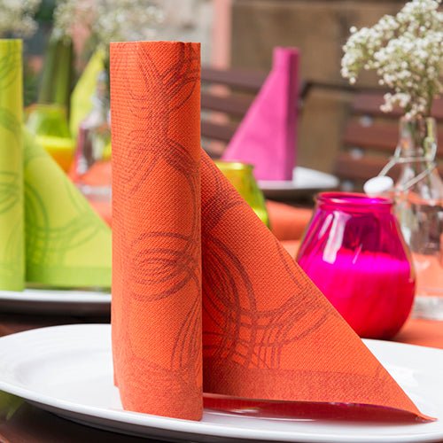A neatly folded orange PAPSTAR 50 "ROYAL Collection" napkins, 1/4-fold, 40 x 40 cm. A peach-colored napkin stands upright on a white plate, featuring colorful ROYAL Collection designs and blurred flowers in the background.