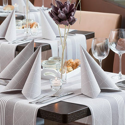 A restaurant table set with PAPSTAR GmbH 50 napkins "ROYAL Collection" 1/4-fold, 40 x 40 cm, brown, folded into triangles, with wine glasses, cutlery, a bread basket, a purple artificial flower centerpiece, and a burning tealight.