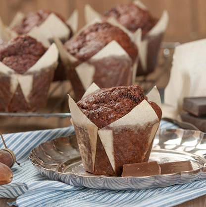 A chocolate muffin in the PAPSTAR baking mold "Tulip" Ø 5 cm, white from PAPSTAR GmbH sits on a silver plate with two pieces of chocolate; more muffins in decorative wrappers cool on a rack over a blue striped cloth.