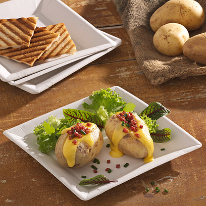 Two baked potatoes with yellow cheese sauce, bacon bits, and chives are served on a PAPSTAR square paper plate 22.5 cm in the "pure" design by PAPSTAR GmbH. Leafy greens accompany the dish, with grilled bread and whole potatoes in the background.