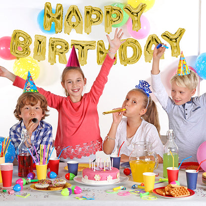 Vier Kinder mit Partyhüten sitzen an einem festlich gedeckten Tisch mit Kuchen und Getränken. An der Wand sorgt das Folienluftballon-Set „Happy Birthday“ der PAPSTAR GmbH für goldenen Glanz bei der Geburtstagsfeier.