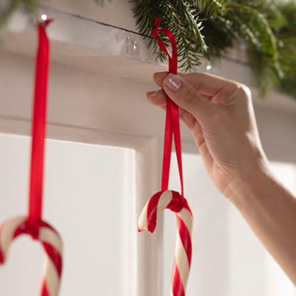 A hand hangs a candy cane with a red ribbon on a Command™ Mini Hook, transparent, with transparent strips (3M Deutschland GmbH), which is attached under a shelf decorated with evergreen branches. Another candy cane is already hanging on the hook next to it.