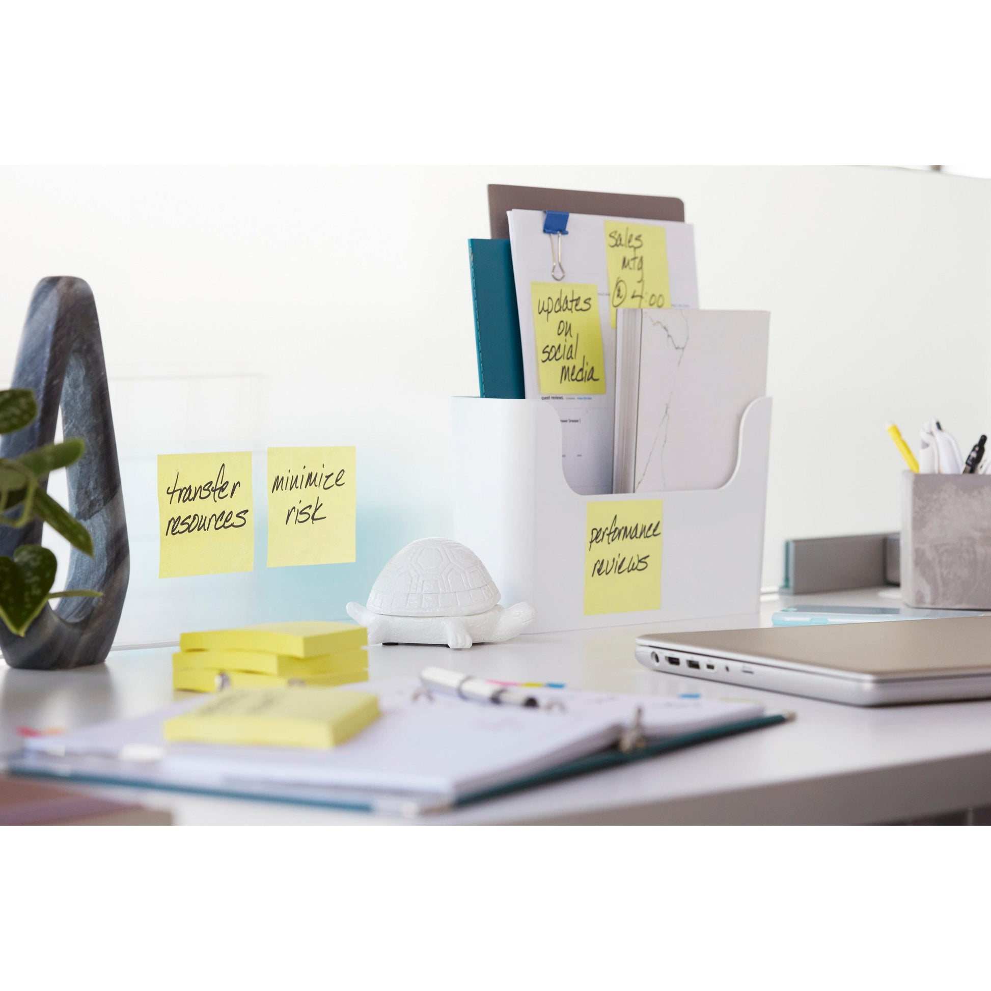 Ein aufgeräumter Bürotisch mit Post-it® Super Sticky Notes (76x127 mm, 90 Blatt, 100% PEFC) von 3M Deutschland GmbH auf einem Glastrenner, dazu Ordner in einem Halter, eine Schildkrötenfigur, Büromaterial, ein geschlossener Laptop und ein offenes Notizbuch mit Stift.