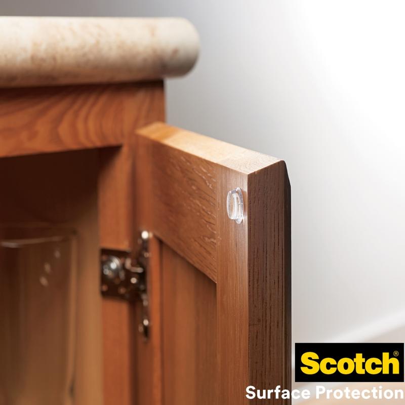 A Scotch® shock-absorbing elastic bumper, self-adhesive, transparent from 3M Germany GmbH is attached to the edge of a wooden door to prevent damage. The Scotch Surface Protection logo is visible at the bottom right.