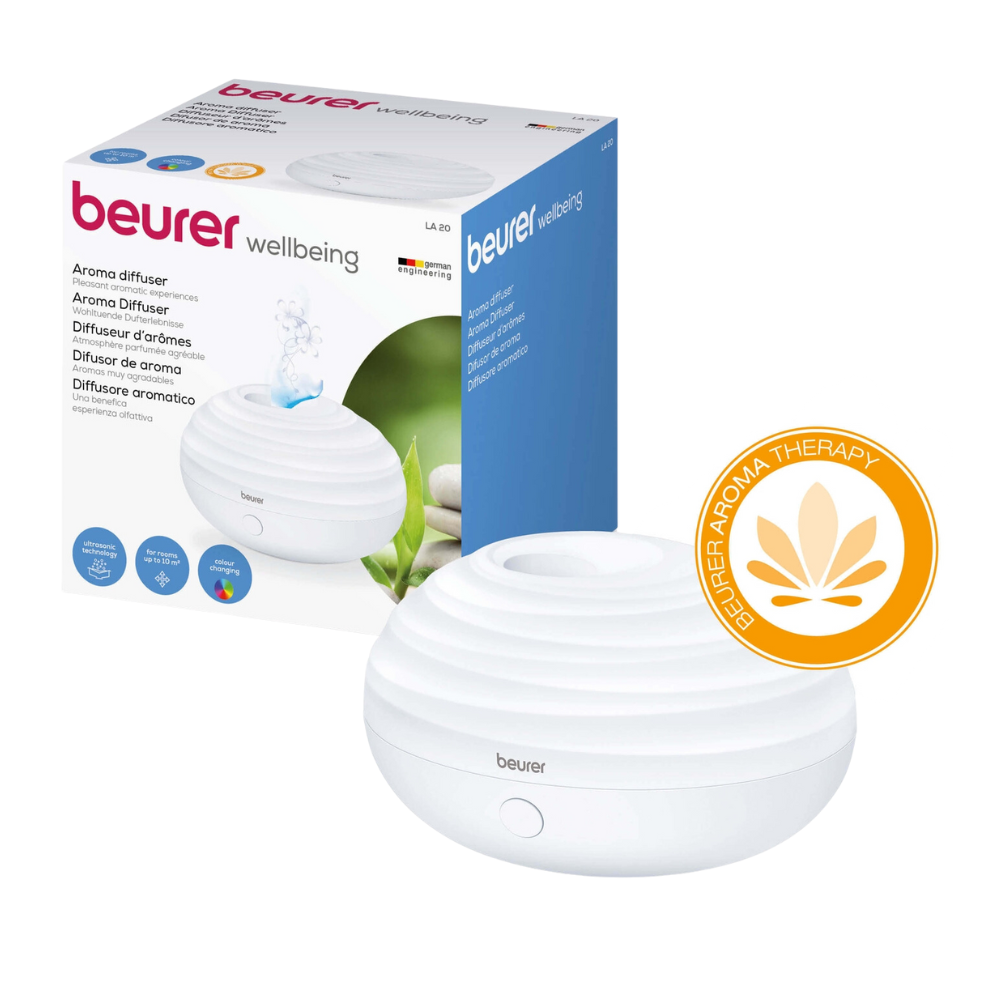 A Beurer Aroma Diffuser LA 20 in white with ultrasonic humidification technology stands next to its packaging. The packaging displays product information and images highlighting the design and LED color light of the diffuser. The image also shows a seal indicating the aromatherapy of Beurer GmbH.
