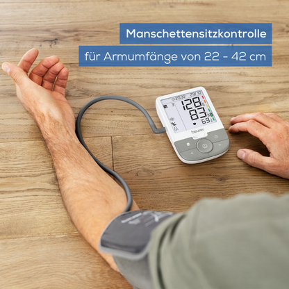 A user is working with the Beurer GmbH discontinued item: upper arm blood pressure monitor BM 53 on a light wooden surface. The display shows "Cuff fit control for arm circumferences of 22-42 cm" and points out arrhythmia detection and stroke prevention.
