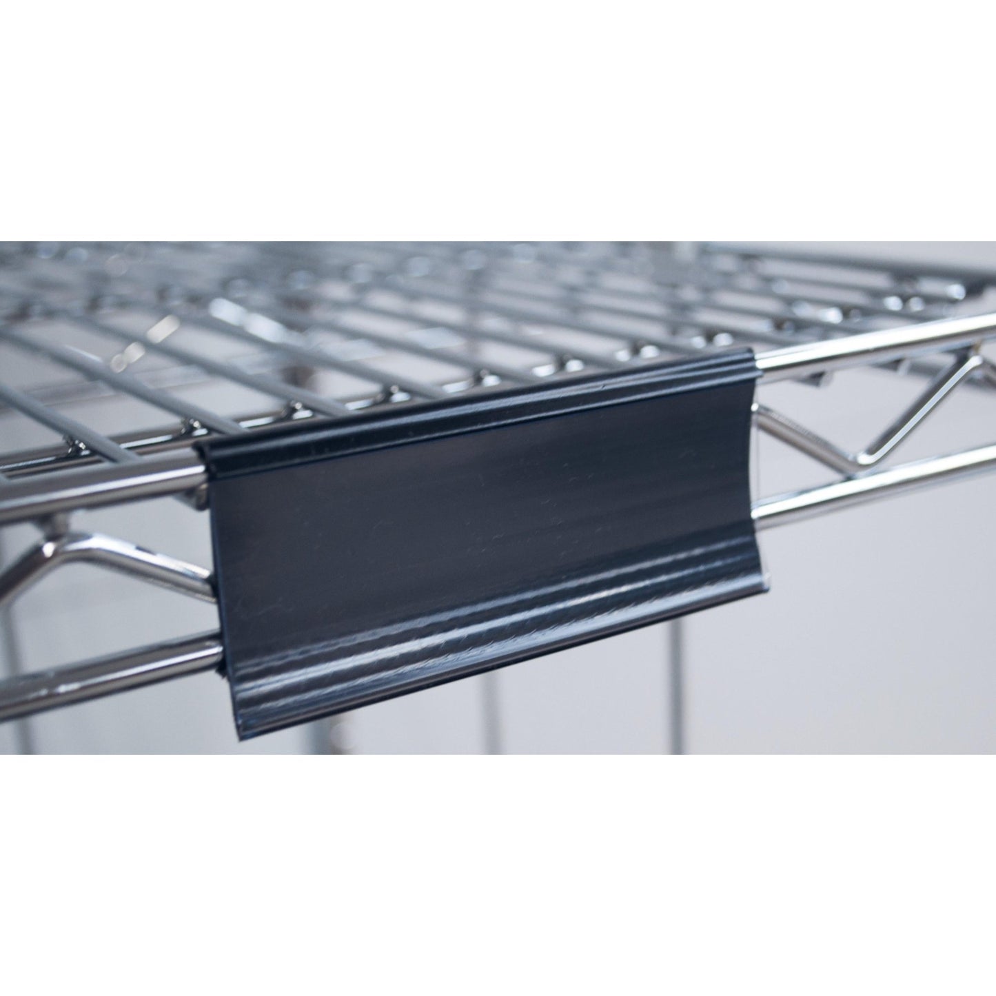 Close-up of a Novocal label frame with paper tags (approx. 100 x 44 mm, 1 piece) attached to a metal wire shelf, with the black plastic label holder on the front edge; the background is blurred.