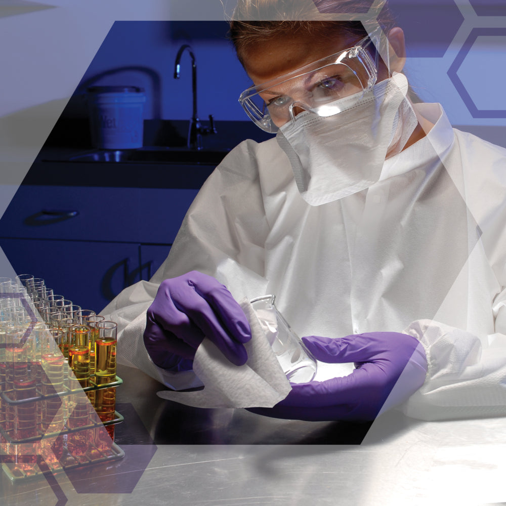 In einem Labor wischt jemand, der eine Schutzbrille, eine Maske und lila Handschuhe trägt, ein Becherglas mit Kimtech® Pure Wischtüchern (Kimberly-Clark GmbH) ab, umgeben von Reagenzgläsern mit farbigen Flüssigkeiten und verschiedenen Laborgeräten.