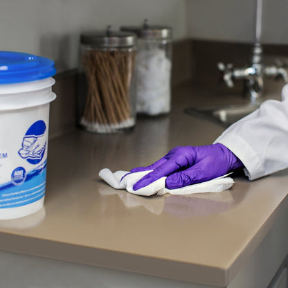 Eine Person mit lila Handschuhen wischt mit WypAll® Wettask™ Wischtüchern der Kimberly-Clark GmbH (6 Rollen x 90 Blatt) eine beigefarbene Arbeitsplatte in der Nähe des Waschbeckens ab, während im Hintergrund Gläser mit Wattestäbchen und -bällen zu sehen sind.