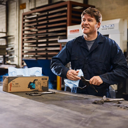 A man in a dark overall stands at a workbench and cleans a tool with a blue WypAll® X60 General Clean towel from Kimberly-Clark GmbH. A box labeled "Central Dispensing / Blue - 1 Roll x 150 Sheets" stands next to several metal tools on the workbench.
